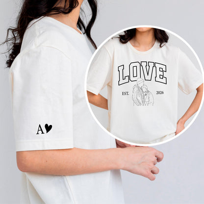 May include: Cream-colored t-shirt featuring the word "LOVE" in bold, black, collegiate-style lettering above a line drawing of two people embracing. The shirt also has the text "EST. 2026". The left sleeve has the letter "A" and a heart.