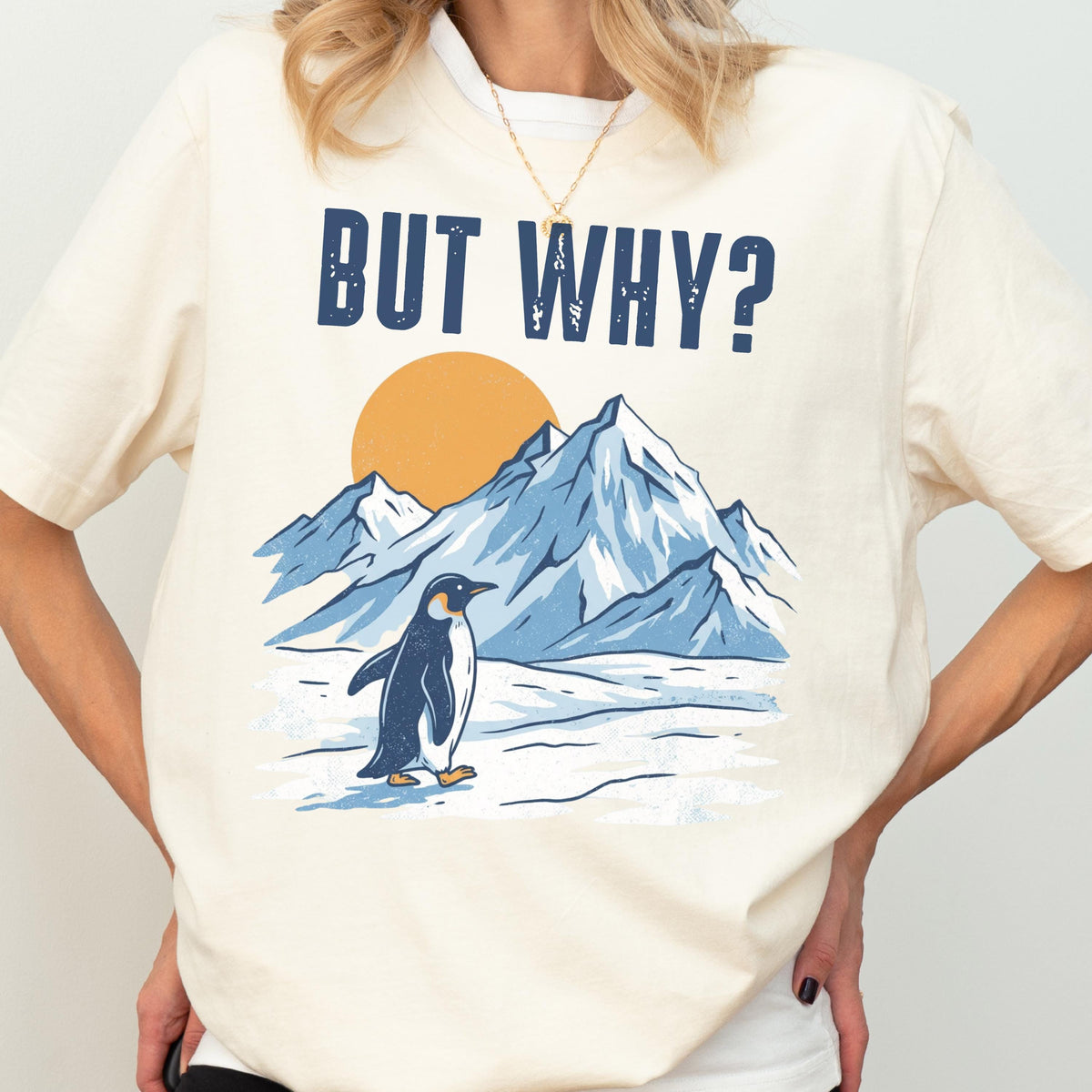 May include: Cream-colored t-shirt featuring a graphic of a penguin standing in front of snow-covered mountains and a yellow sun. The text "BUT WHY?" is printed above the graphic in navy blue.