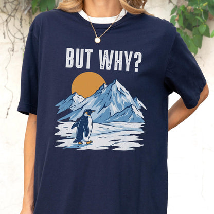May include: Navy blue t-shirt with a white collar featuring a graphic of a penguin, mountains, and a sun. The text "BUT WHY?" is printed above the graphic in white block letters. The shirt is a casual style.