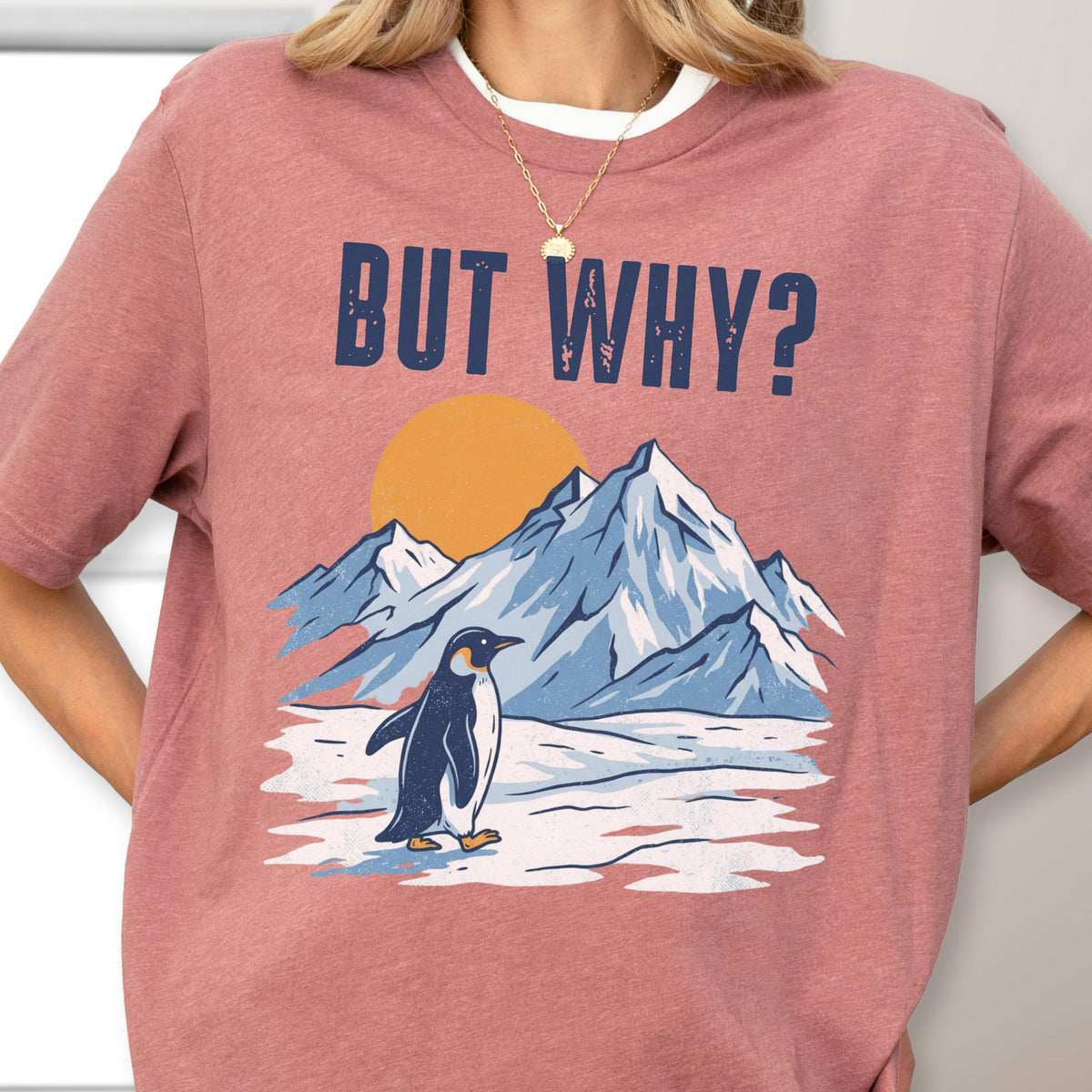 May include: A dusty rose t-shirt with the phrase "BUT WHY?" in navy blue block letters. Below the text is a graphic of a penguin standing in front of snow-covered mountains and a setting sun. The penguin is black and white.