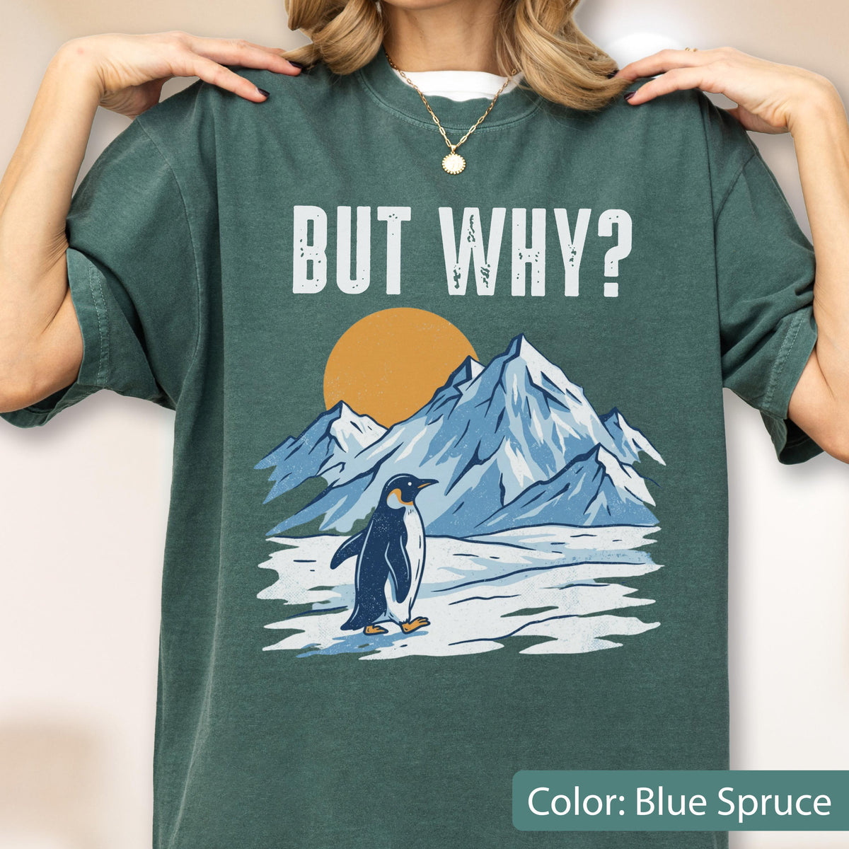 May include: Blue spruce-colored t-shirt with the text "BUT WHY?" above a graphic of a penguin standing in front of snow-covered mountains and a setting sun. The shirt is a solid color.