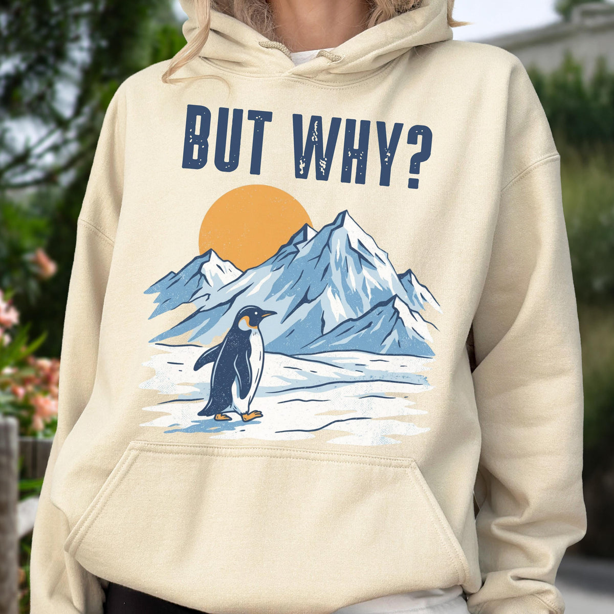 May include: A beige hoodie with a graphic featuring a penguin standing in front of snow-covered mountains and a setting sun. The text "BUT WHY?" is printed above the scene in navy blue block letters. The hoodie has a drawstring hood and a front pocket.