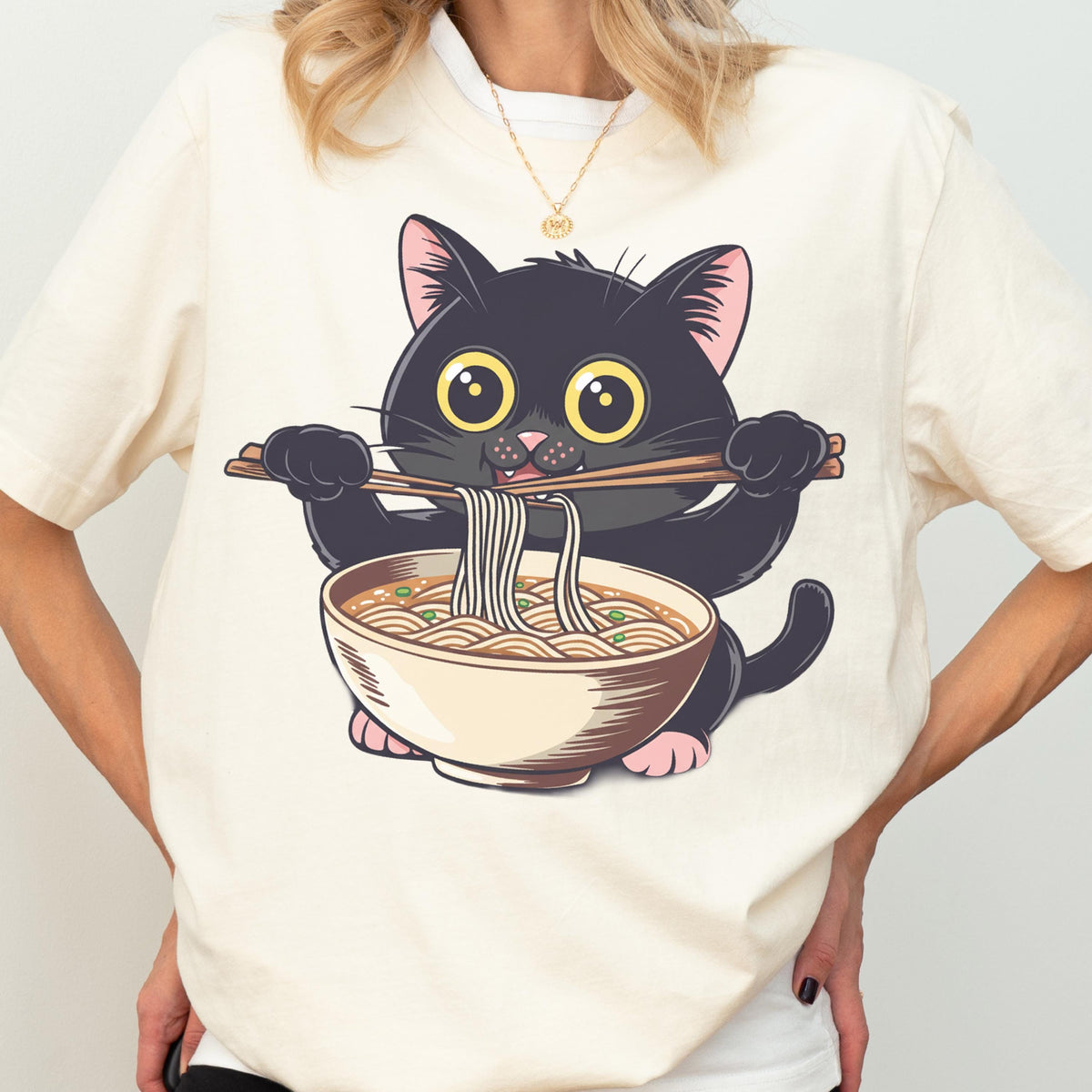May include: Cream-colored t-shirt featuring a cartoon black cat with large yellow eyes, holding chopsticks with noodles over a bowl of ramen. The cat has pink paws and ears. The design is centered on the shirt.