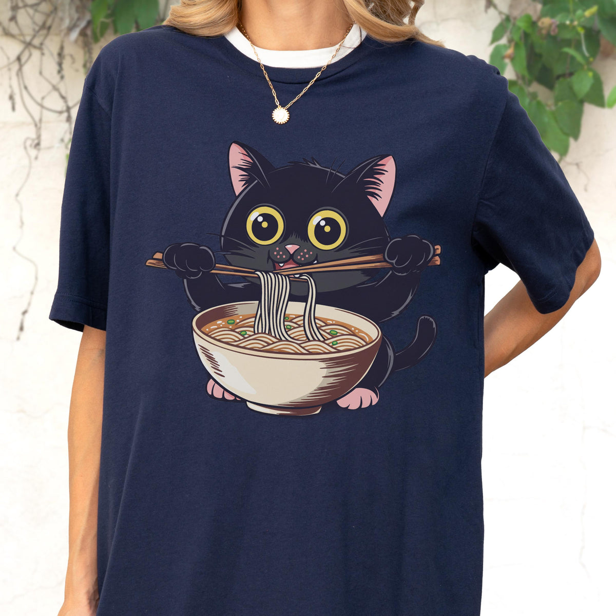 May include: Navy blue t-shirt featuring a cartoon black cat with large yellow eyes, holding chopsticks and a bowl of noodles. The cat is depicted with pink ears and paws. The shirt has a white collar.
