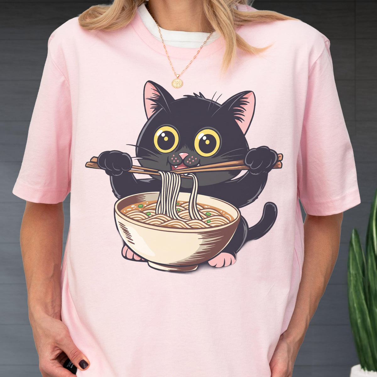 May include: Pink t-shirt featuring a cartoon black cat holding chopsticks and eating ramen noodles from a bowl. The cat has large yellow eyes and pink paws. The shirt is a light pink color.