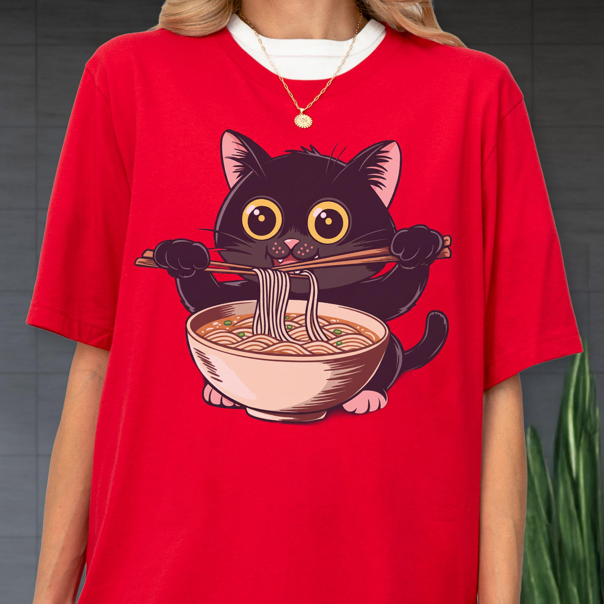 May include: Red t-shirt featuring a cartoon black cat eating ramen noodles with chopsticks. The cat has large yellow eyes and is holding a bowl of noodles. The shirt has a white collar and is a casual style.
