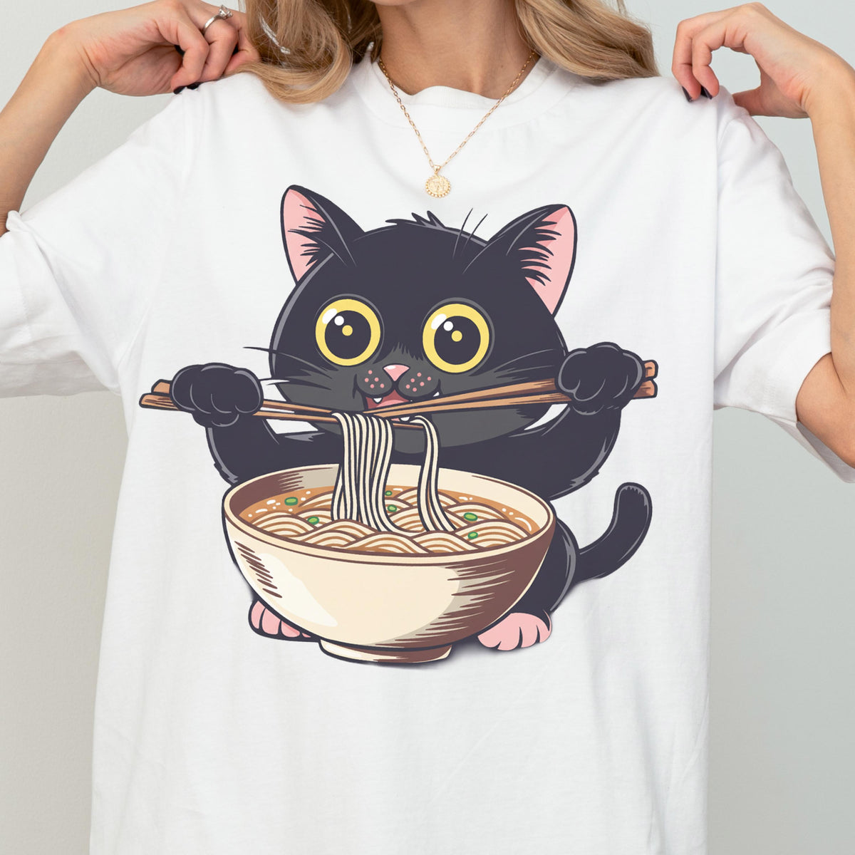 May include: White t-shirt featuring a cartoon black cat with large yellow eyes, holding chopsticks and eating ramen from a bowl. The cat is black with pink accents on its paws and ears. The ramen bowl is beige with a brown broth.