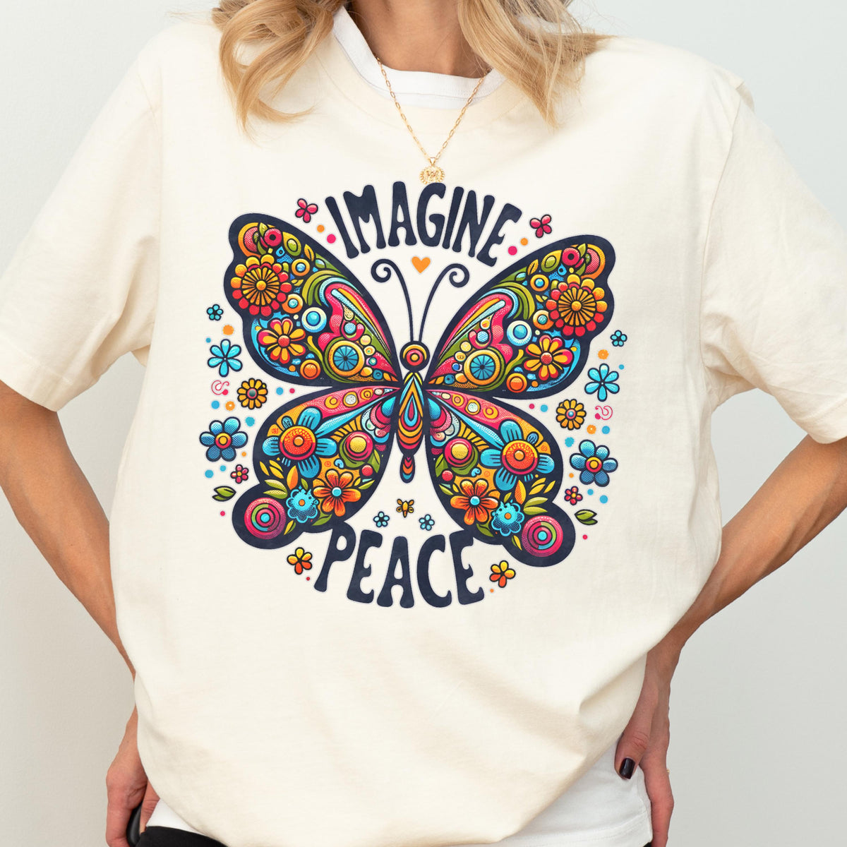 May include: A cream-colored t-shirt featuring a large, colorful butterfly graphic with intricate floral patterns in shades of red, orange, yellow, and blue. The text "IMAGINE" is printed in a dark blue font above the butterfly, and "PEACE" below.