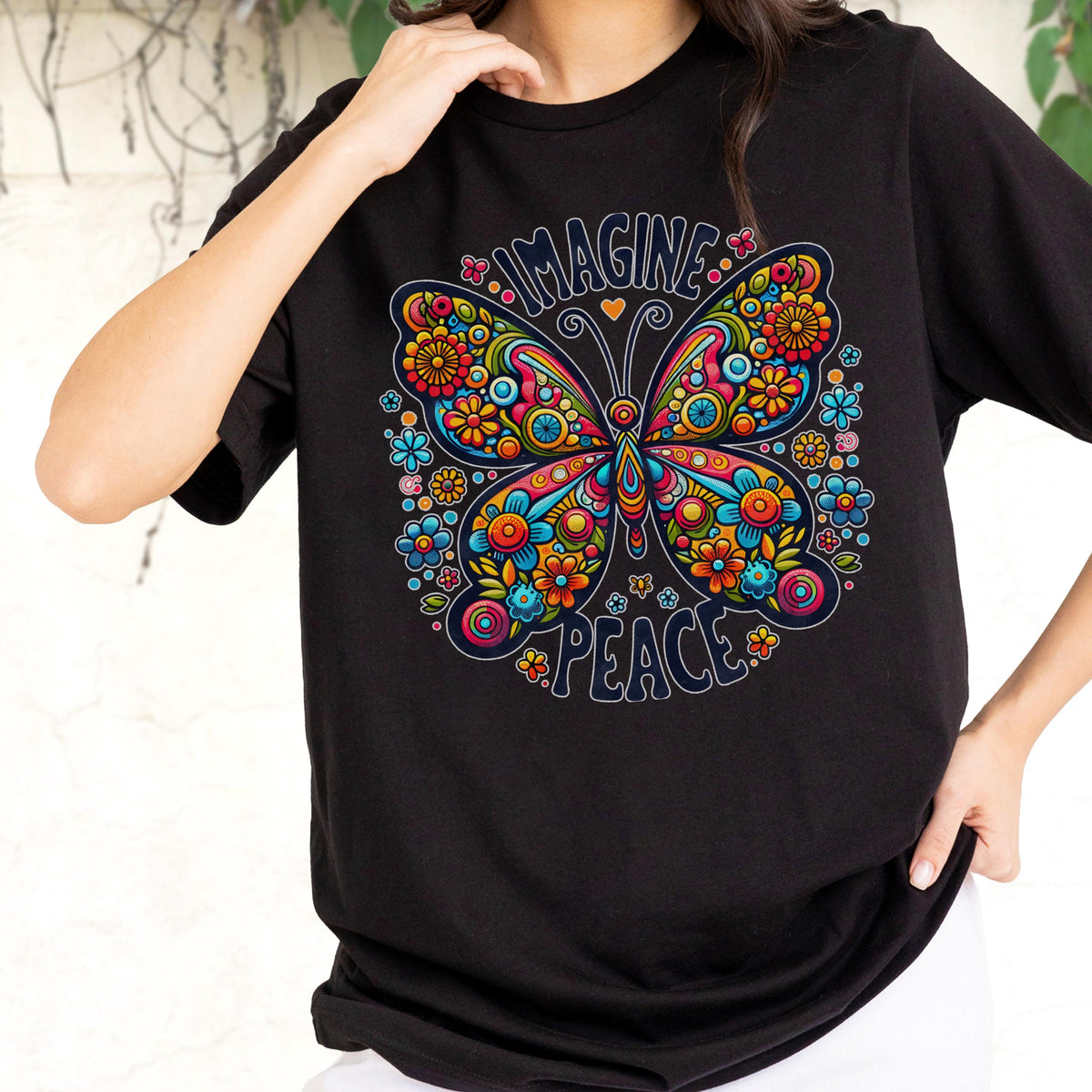 May include: A black short-sleeved t-shirt featuring a large, colorful butterfly graphic composed of intricate floral patterns in shades of red, orange, yellow, and blue. The text "IMAGINE" is arched above the butterfly, and "PEACE" is below it.