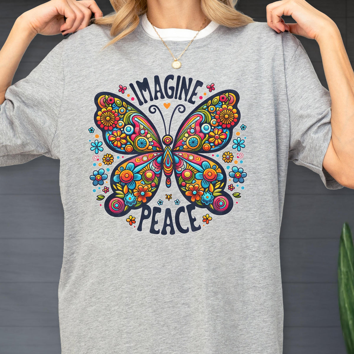 May include: A heather gray t-shirt featuring a large, colorful butterfly graphic with intricate floral patterns in shades of red, orange, yellow, and blue. The text "IMAGINE" is arched above the butterfly, and "PEACE" is printed below it in dark blue.