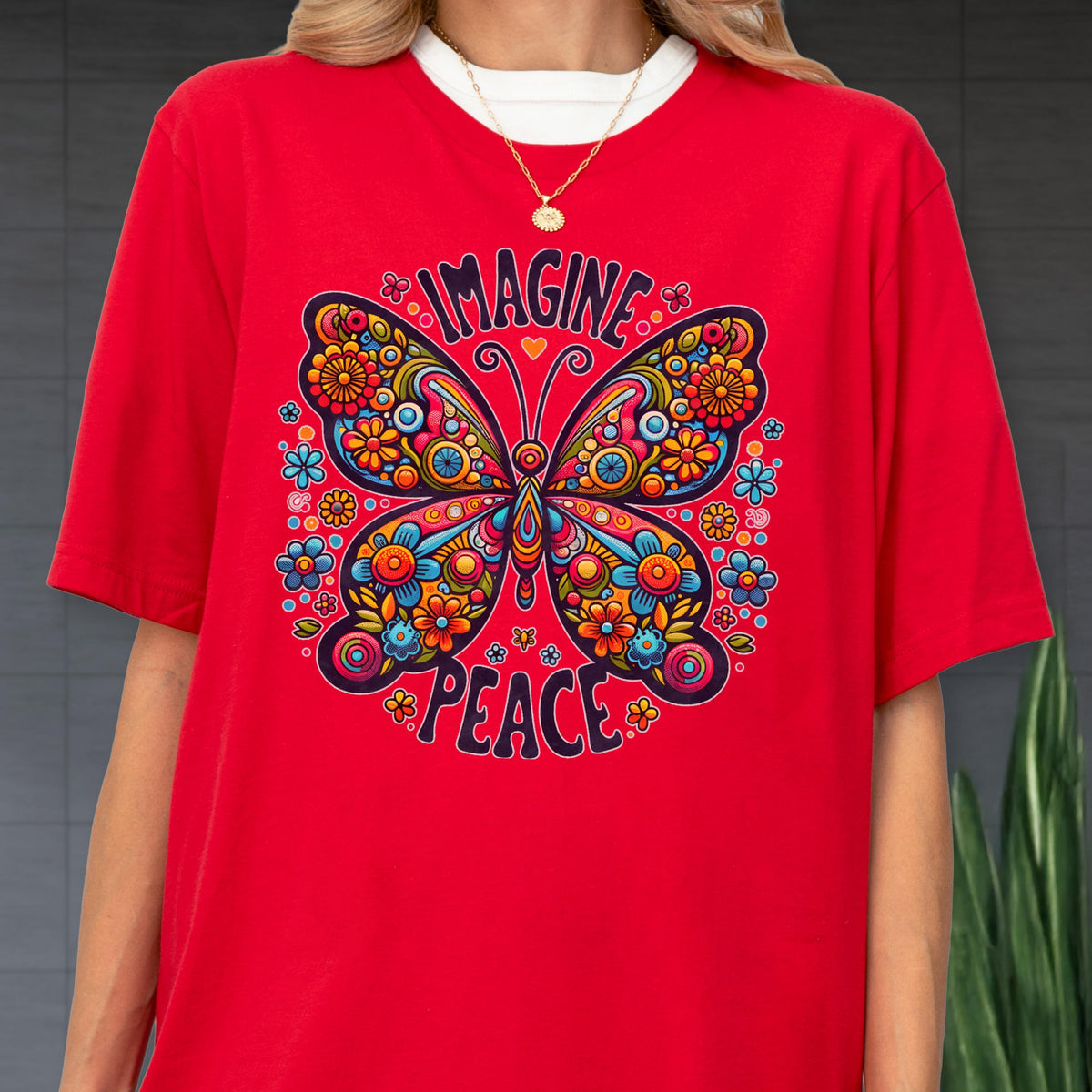 May include: A bright red short-sleeved t-shirt featuring a large, colorful butterfly graphic with intricate floral patterns in shades of blue, orange, yellow, and pink. The text "IMAGINE" is arched above the butterfly and "PEACE" is below it.