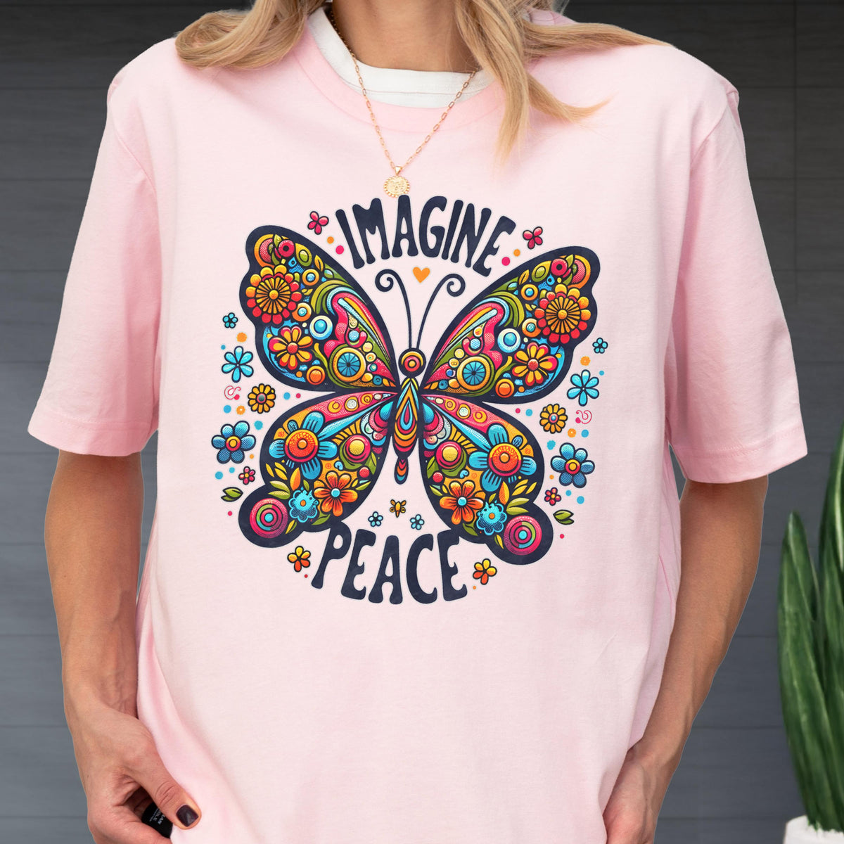 May include: A light pink t-shirt featuring a large, colorful butterfly graphic with intricate floral patterns in shades of blue, orange, yellow, and red. The text "IMAGINE" is arched above the butterfly, and "PEACE" is printed below in dark blue.