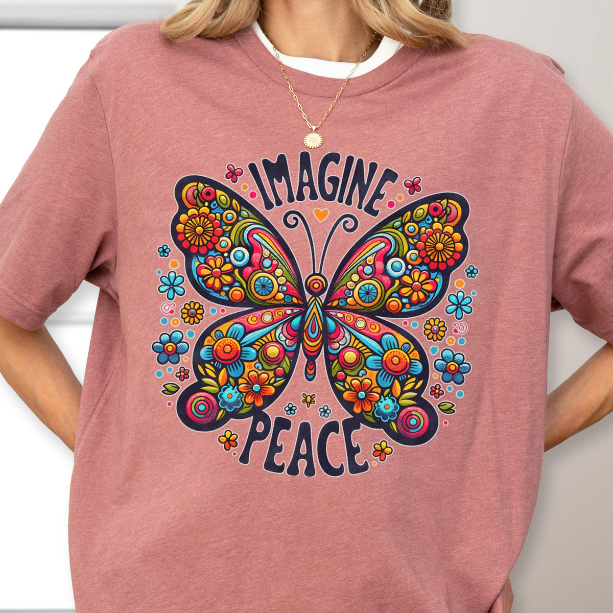 May include: A dusty rose short-sleeve t-shirt featuring a large, colorful butterfly graphic with intricate floral patterns in shades of blue, orange, yellow, and pink. The text "IMAGINE" is arched above the butterfly, and "PEACE" is below it.