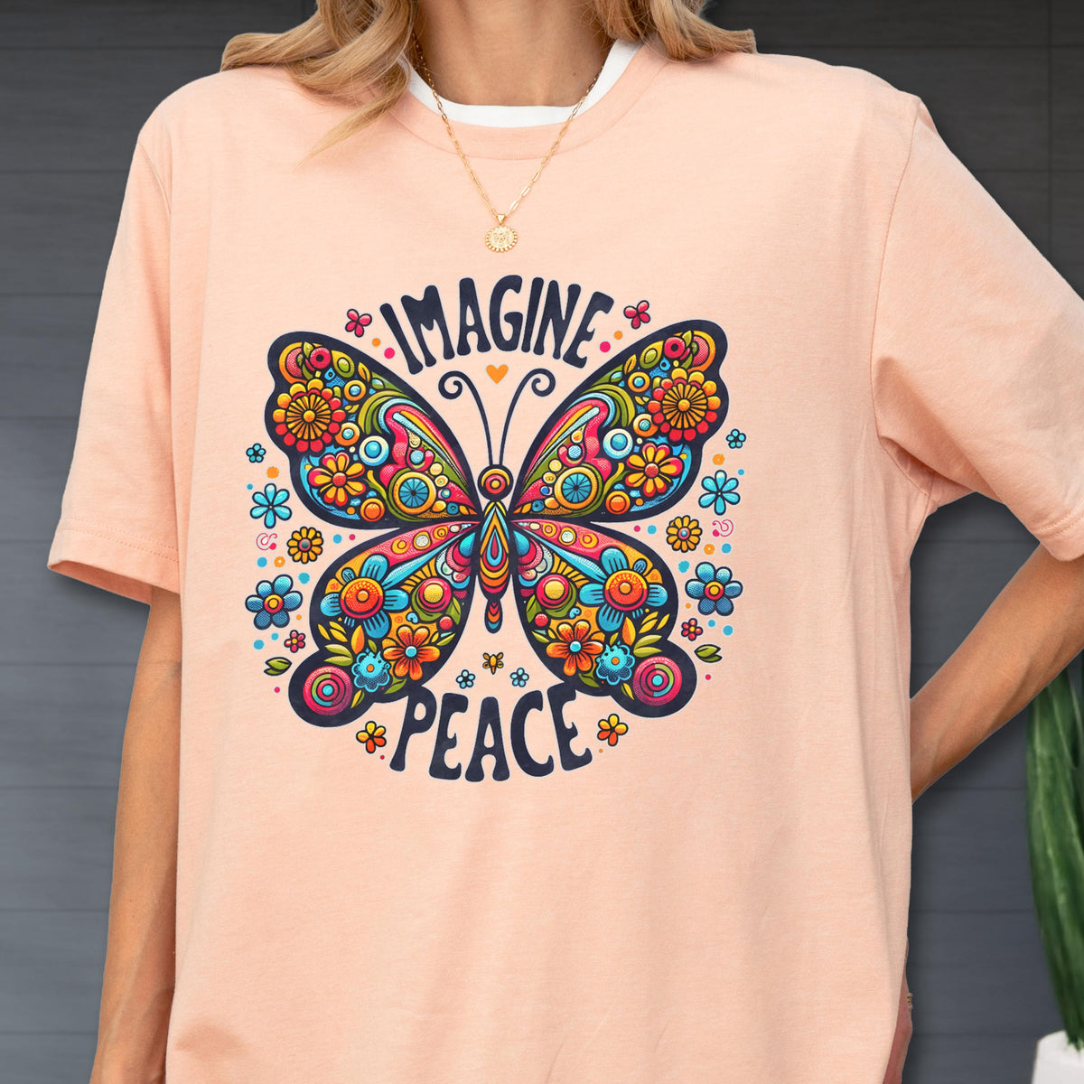 May include: A peach-colored t-shirt featuring a large, colorful butterfly graphic with intricate floral patterns in shades of blue, red, yellow, and orange. The text "IMAGINE" is printed above the butterfly and "PEACE" is printed below it.