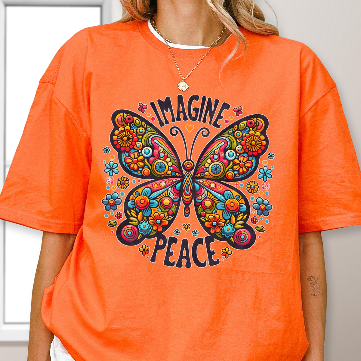 May include: An orange short-sleeved t-shirt featuring a large, colorful butterfly graphic with intricate floral patterns in shades of red, yellow, blue, and pink. The text "IMAGINE" is printed above the butterfly and "PEACE" is printed below it.