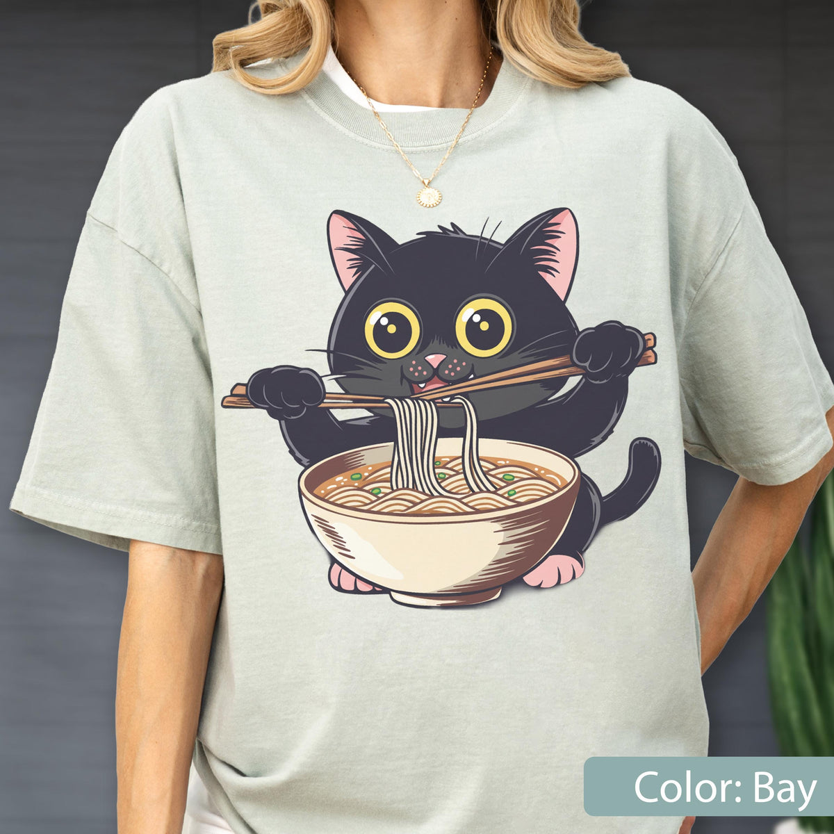May include: A light green t-shirt featuring a cartoon black cat eating ramen noodles with chopsticks. The cat has large yellow eyes and pink ears. The ramen bowl is beige with brown details. The text "Color: Bay" is at the bottom.