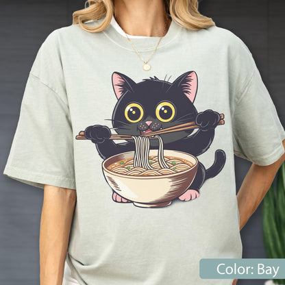 May include: A light green t-shirt featuring a cartoon black cat eating ramen noodles with chopsticks. The cat has large yellow eyes and pink ears. The ramen bowl is beige with brown details. The text "Color: Bay" is at the bottom.