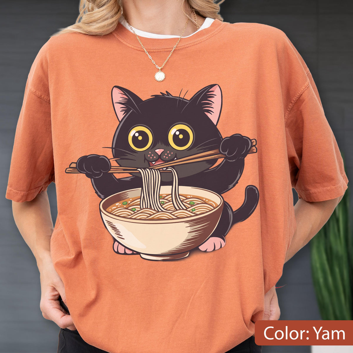 May include: Orange t-shirt featuring a cartoon black cat holding chopsticks and eating ramen noodles from a bowl. The cat has large yellow eyes and pink paws. The shirt is a solid color and the text "Color: Yam" is at the bottom.