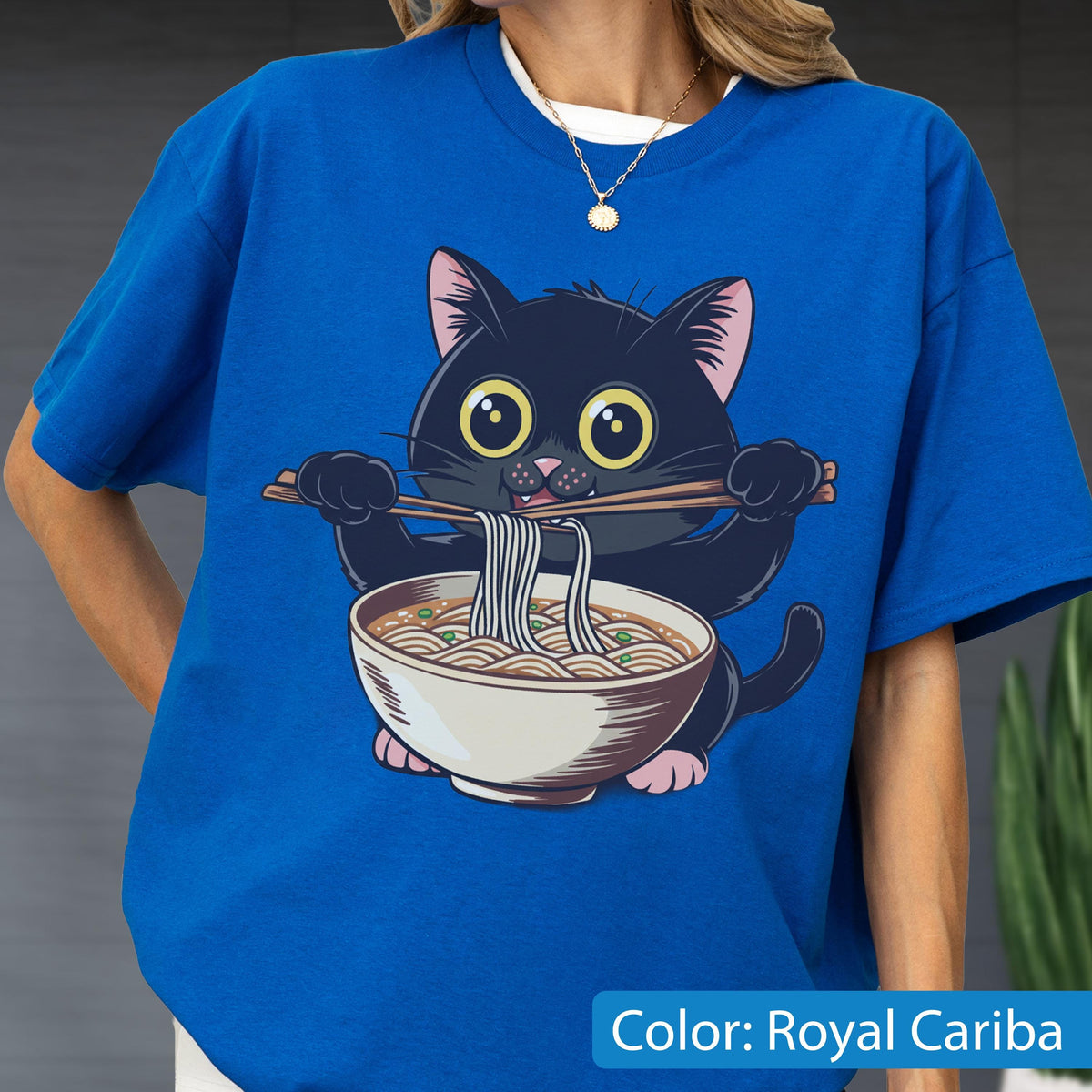 May include: Royal Cariba blue t-shirt featuring a cartoon black cat eating ramen noodles with chopsticks. The cat has large yellow eyes and pink paws. The ramen bowl is white with a brown broth and noodles. The text "Color: Royal Cariba" is at the bottom.