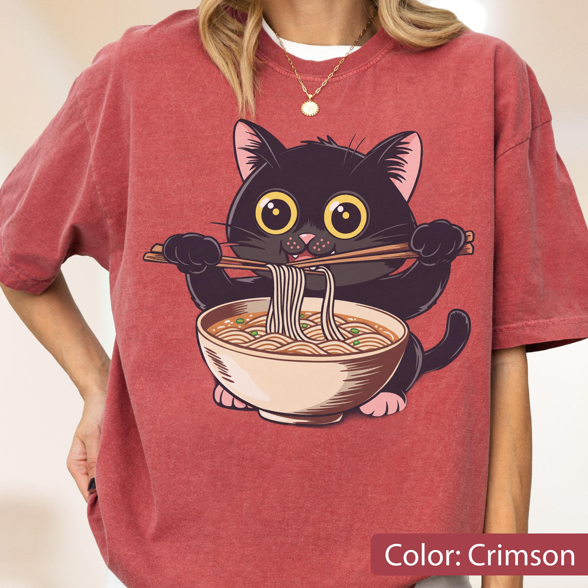 May include: Crimson-colored t-shirt featuring a cartoon black cat holding chopsticks with noodles from a bowl. The cat has large yellow eyes and pink paws. The text "Color: Crimson" is at the bottom.