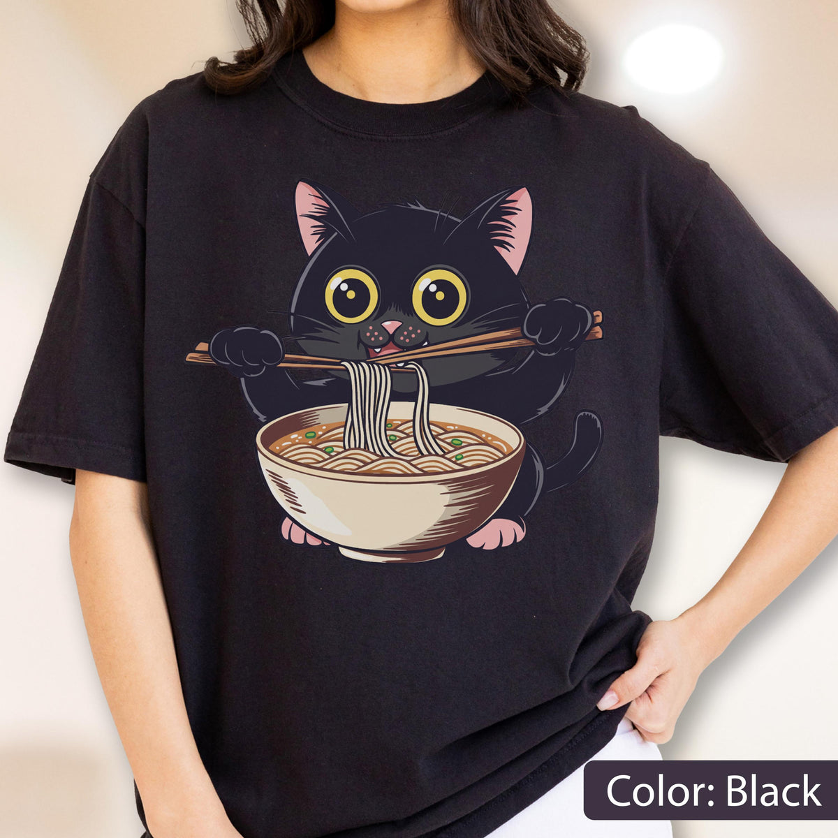 May include: Black t-shirt featuring a cartoon black cat eating ramen noodles with chopsticks. The cat has large yellow eyes and pink ears and paws. The ramen bowl is beige with brown noodles and green garnish. The text "Color: Black" is at the bottom.