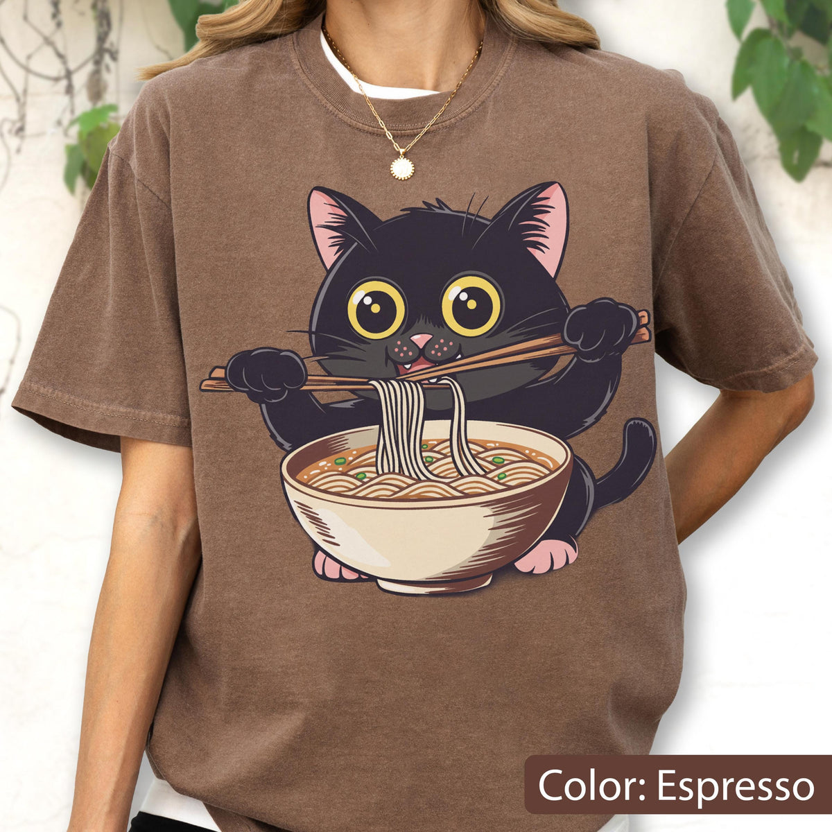 May include: Espresso-colored t-shirt featuring a cartoon black cat enjoying ramen noodles. The cat, with large yellow eyes, holds chopsticks. The ramen bowl is beige with a white rim. The text "Color: Espresso" is at the bottom.