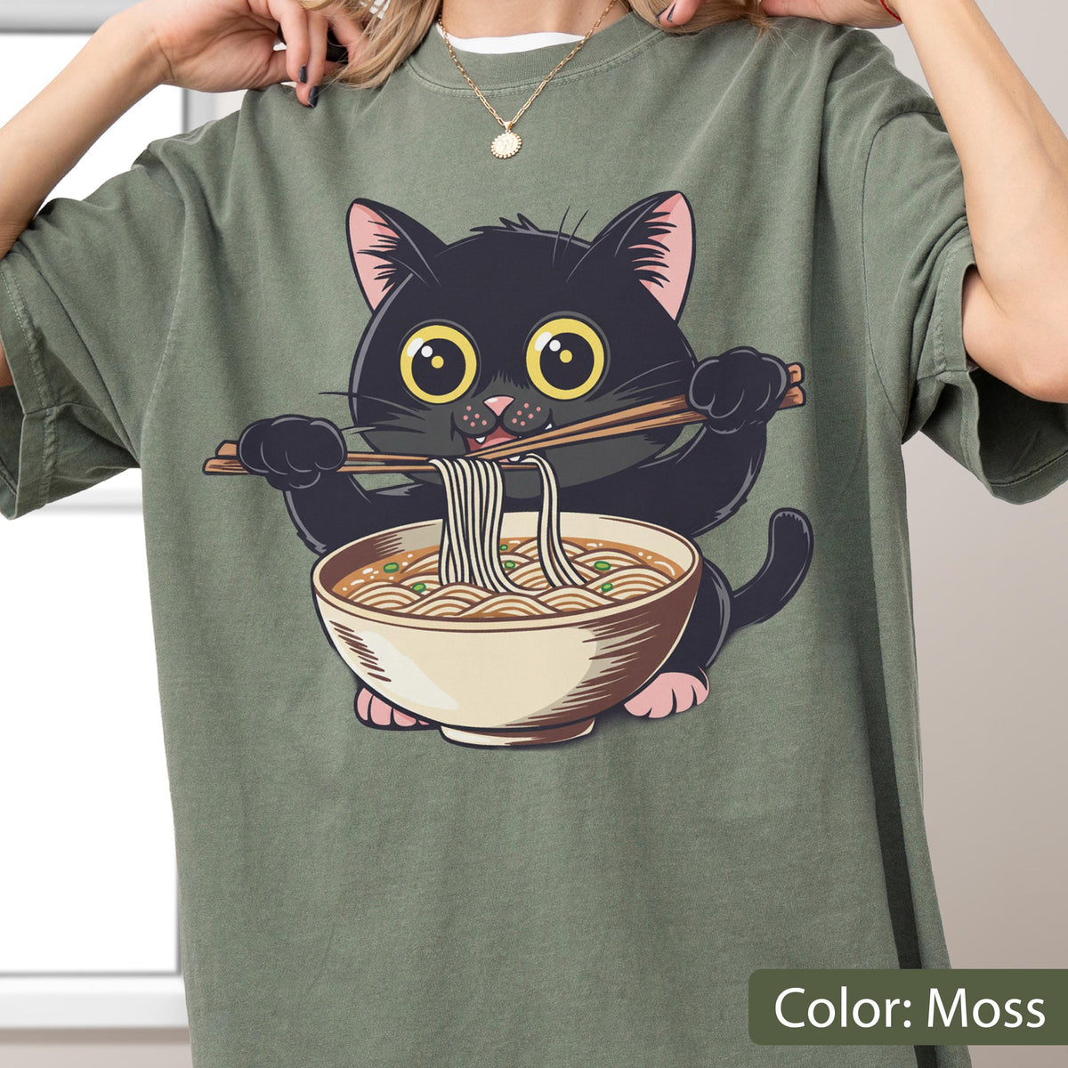 May include: Moss green t-shirt featuring a cartoon black cat eating ramen noodles from a bowl with chopsticks. The cat has large yellow eyes and pink paws. The shirt is a solid color.