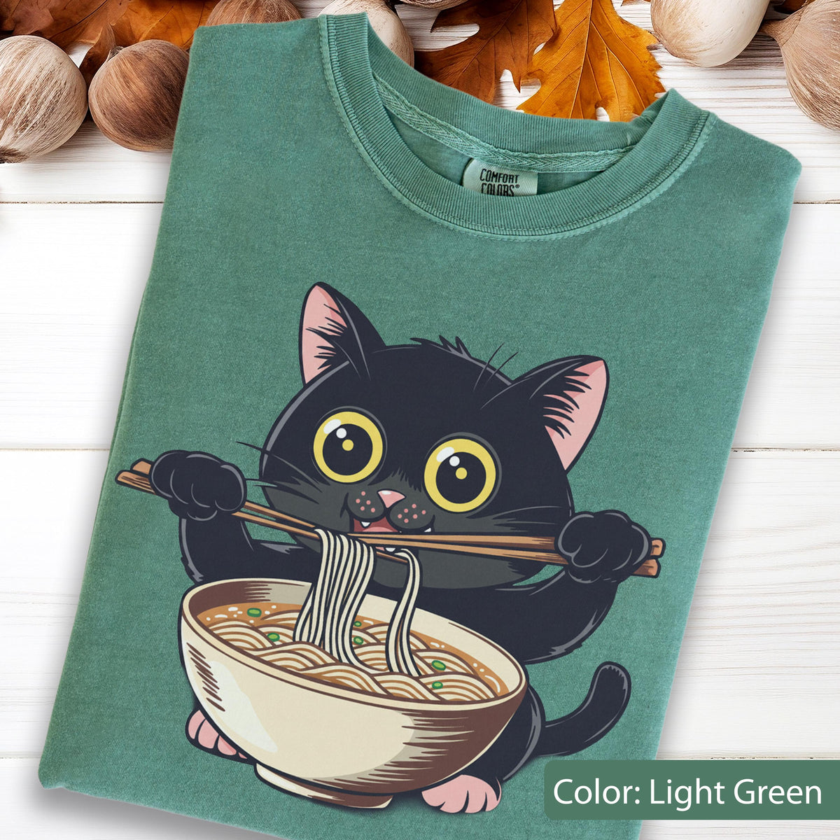 May include: Light green t-shirt featuring a cartoon black cat eating ramen noodles from a bowl with chopsticks. The cat has large yellow eyes and pink ears. The shirt has a crew neck and short sleeves. The text "Color: Light Green" is at the bottom.