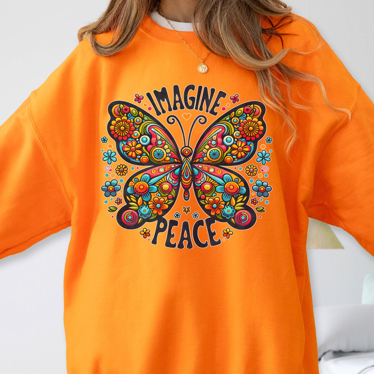 May include: Orange sweatshirt featuring a colorful butterfly design with the words "IMAGINE" and "PEACE". The butterfly has intricate floral patterns in various colors, including blue, yellow, and pink. The sweatshirt is a solid orange color.