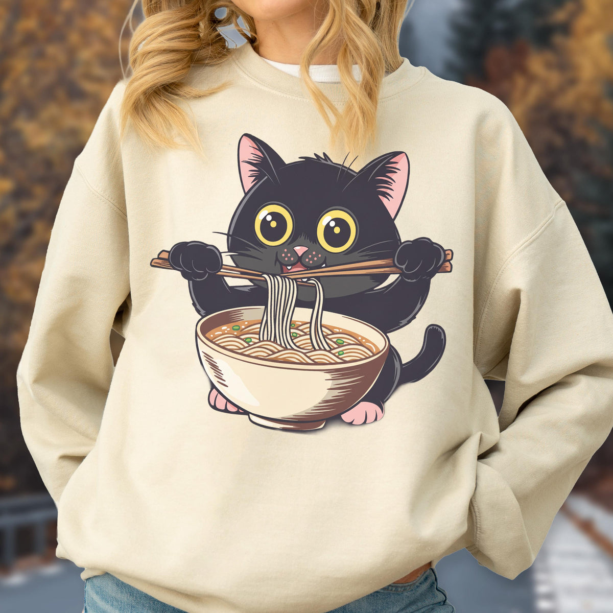 May include: Beige sweatshirt featuring a cartoon black cat holding chopsticks and a bowl of ramen. The cat has large yellow eyes and pink paws. The ramen bowl is beige with brown accents. The sweatshirt is a neutral color.