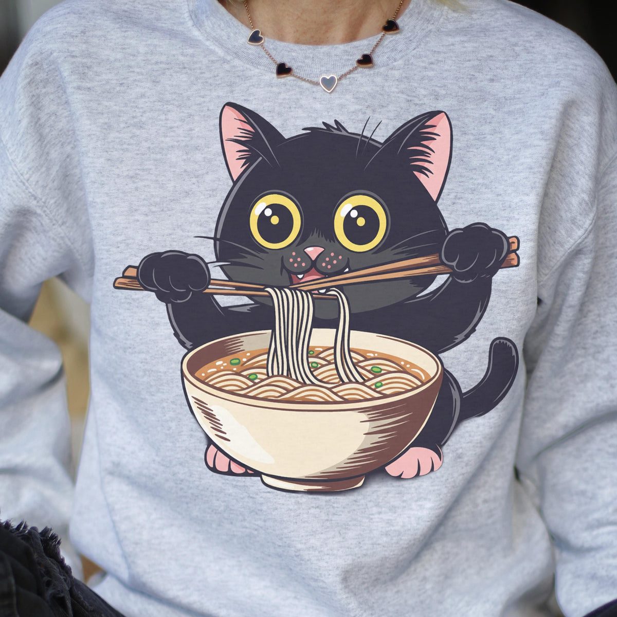 May include: A light gray sweatshirt featuring a cartoon black cat eating ramen noodles with chopsticks. The cat has large yellow eyes and pink inner ears. The ramen bowl is beige with visible noodles and green toppings.