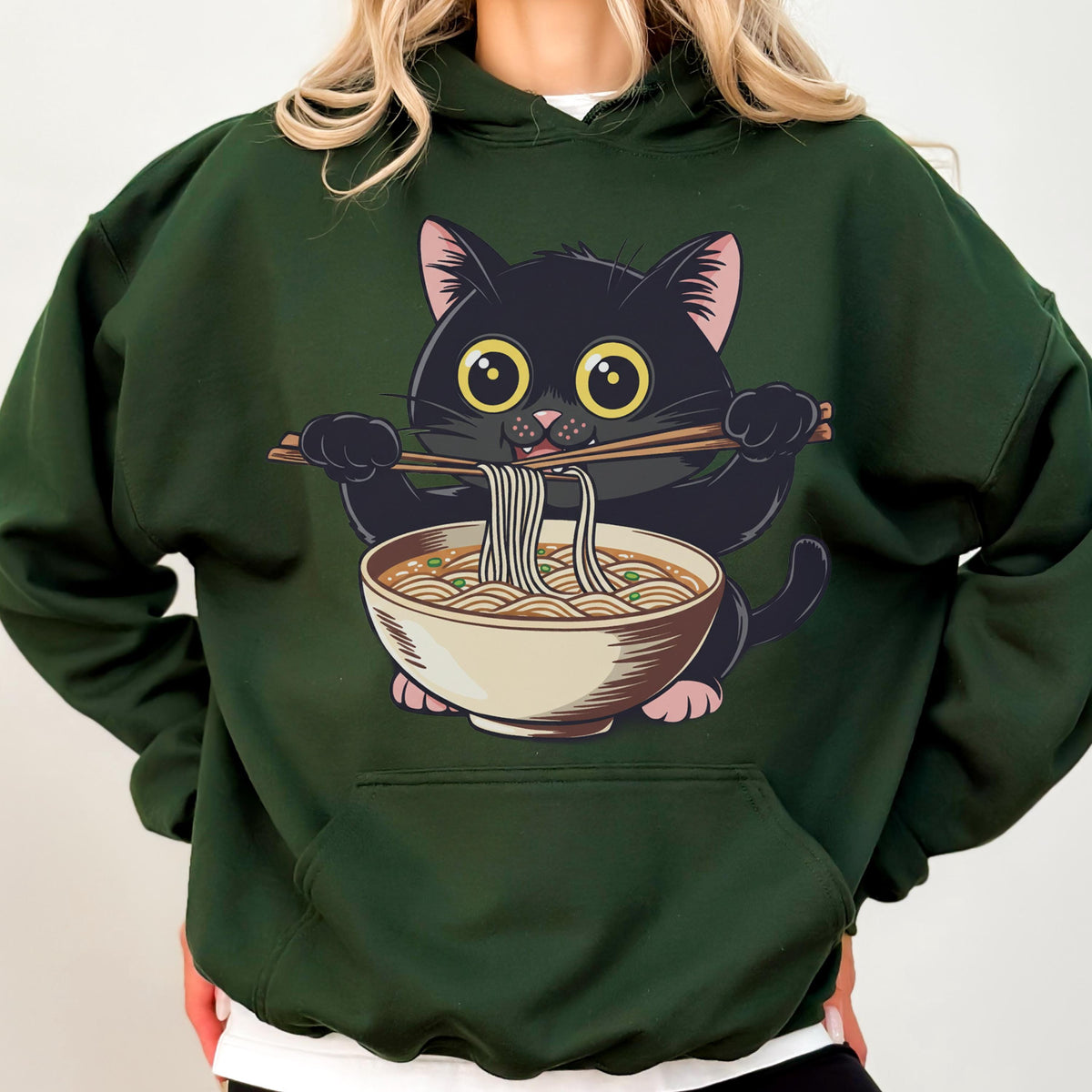 May include: Dark green hoodie featuring a cartoon black cat eating ramen noodles from a beige bowl with chopsticks. The cat has large yellow eyes and pink ears and paws. The hoodie has a front pocket and a drawstring hood.