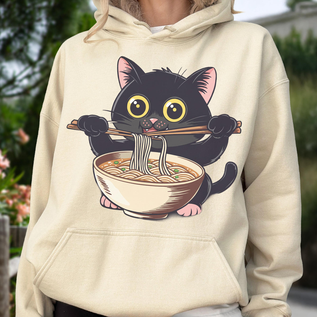 May include: A beige hoodie featuring a cartoon black cat holding chopsticks and a bowl of ramen. The cat has large yellow eyes and pink paws. The ramen bowl is beige with brown noodles and green garnish.