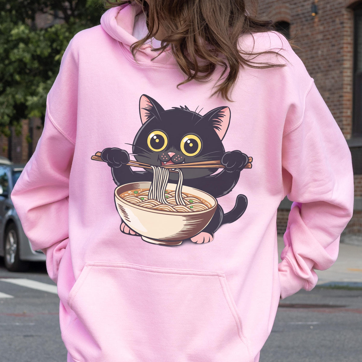 May include: A light pink hoodie featuring a cartoon black cat holding chopsticks with noodles from a bowl. The cat has large yellow eyes and is eating ramen. The hoodie has a front pocket and a hood.