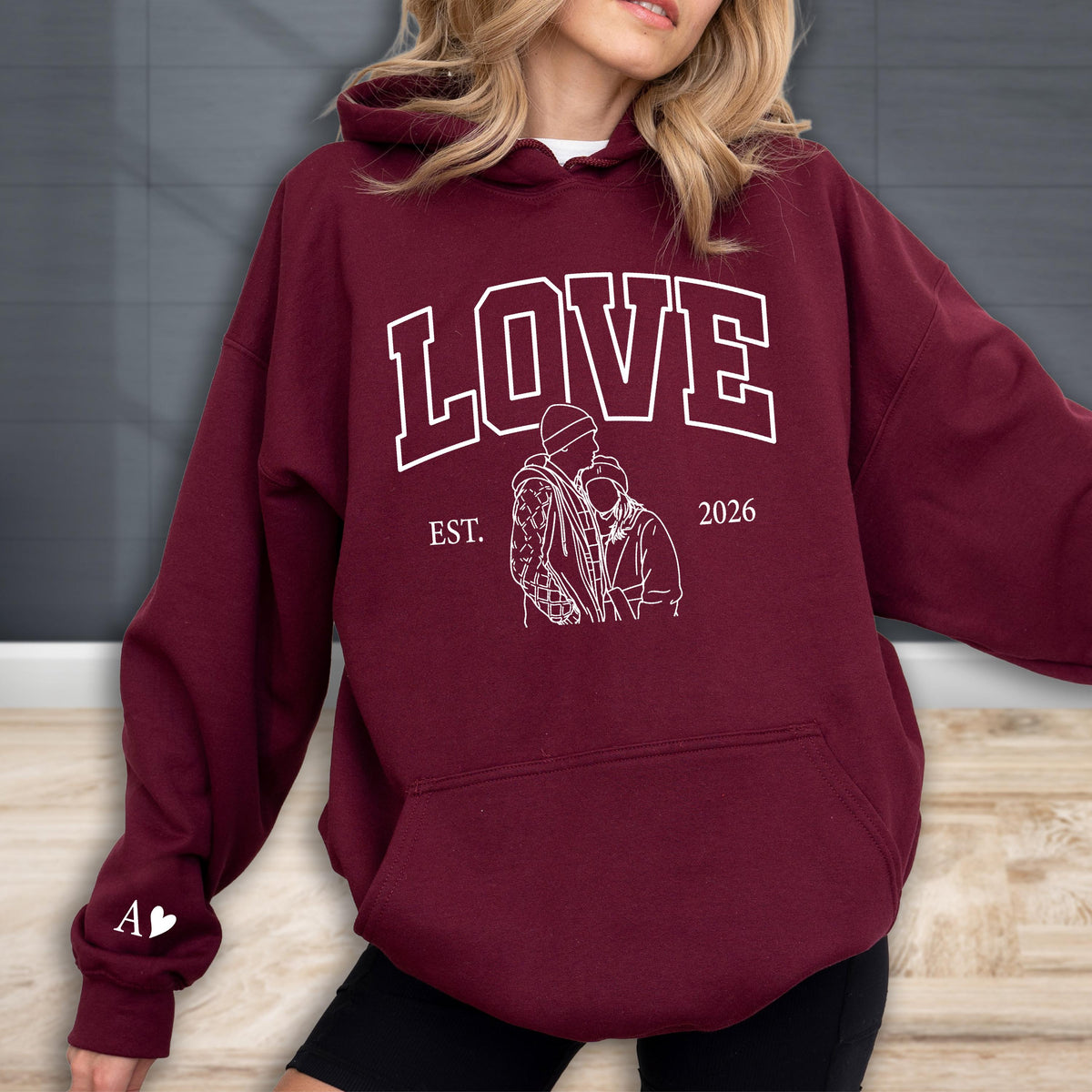 May include: A maroon hoodie with the word "LOVE" in white block letters. Below the word "LOVE" is a white graphic of two people embracing, with the text "EST. 2026" above the graphic. The left sleeve has the letter "A" and a heart.