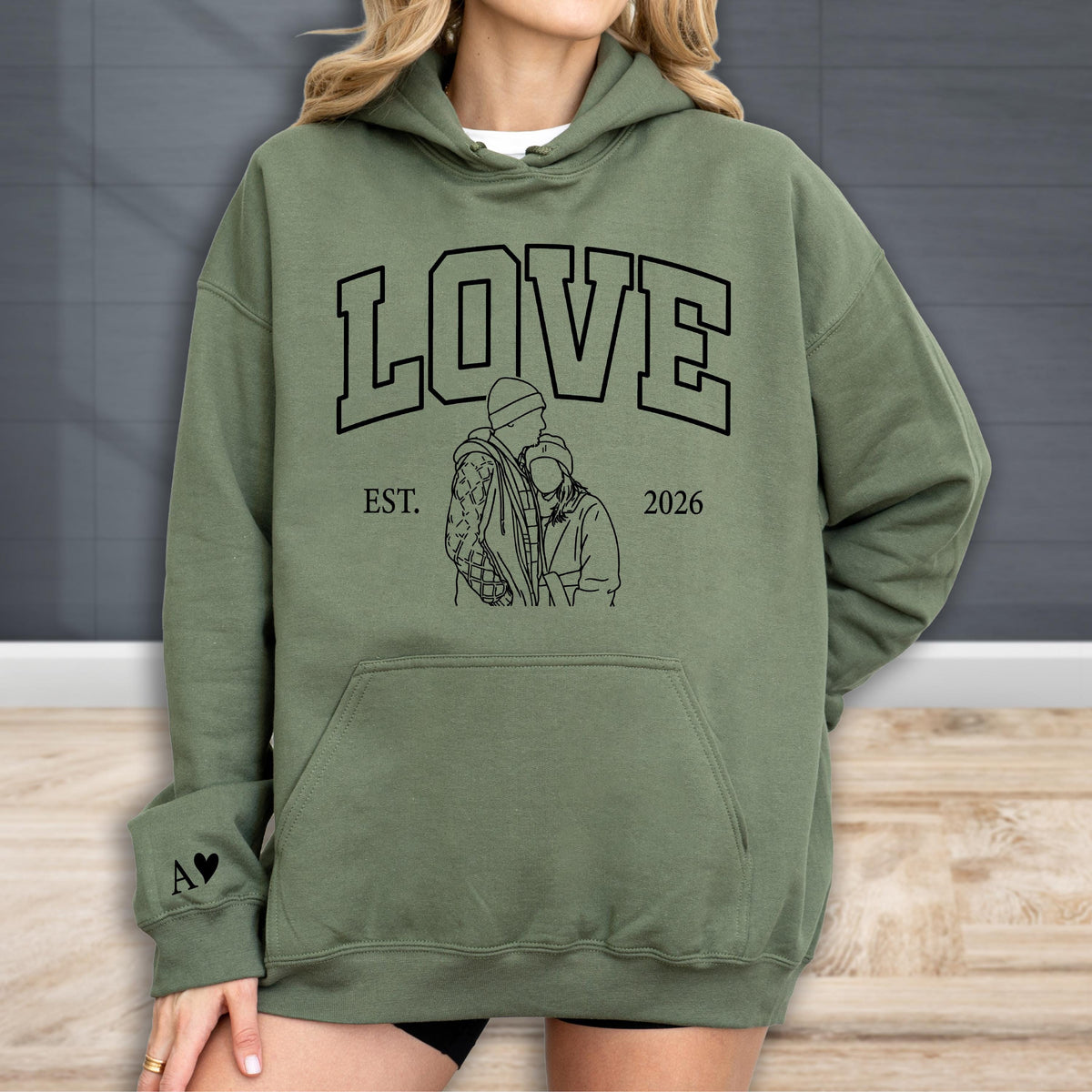 May include: Olive green hoodie with the word "LOVE" in large black block letters. Below the word "LOVE" is a black line drawing of two people embracing, with the text "EST. 2026" below the drawing. A small black heart and the letter "A" are on the left sleeve.