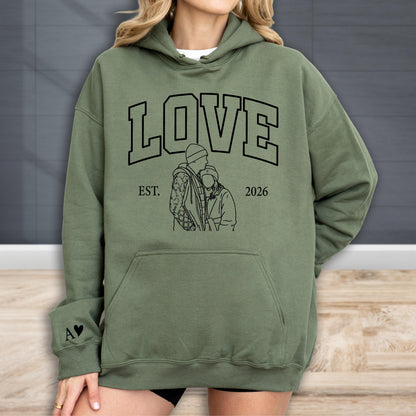 May include: Olive green hoodie with the word "LOVE" in large black block letters. Below the word "LOVE" is a black line drawing of two people embracing, with the text "EST. 2026" below the drawing. A small black heart and the letter "A" are on the left sleeve.