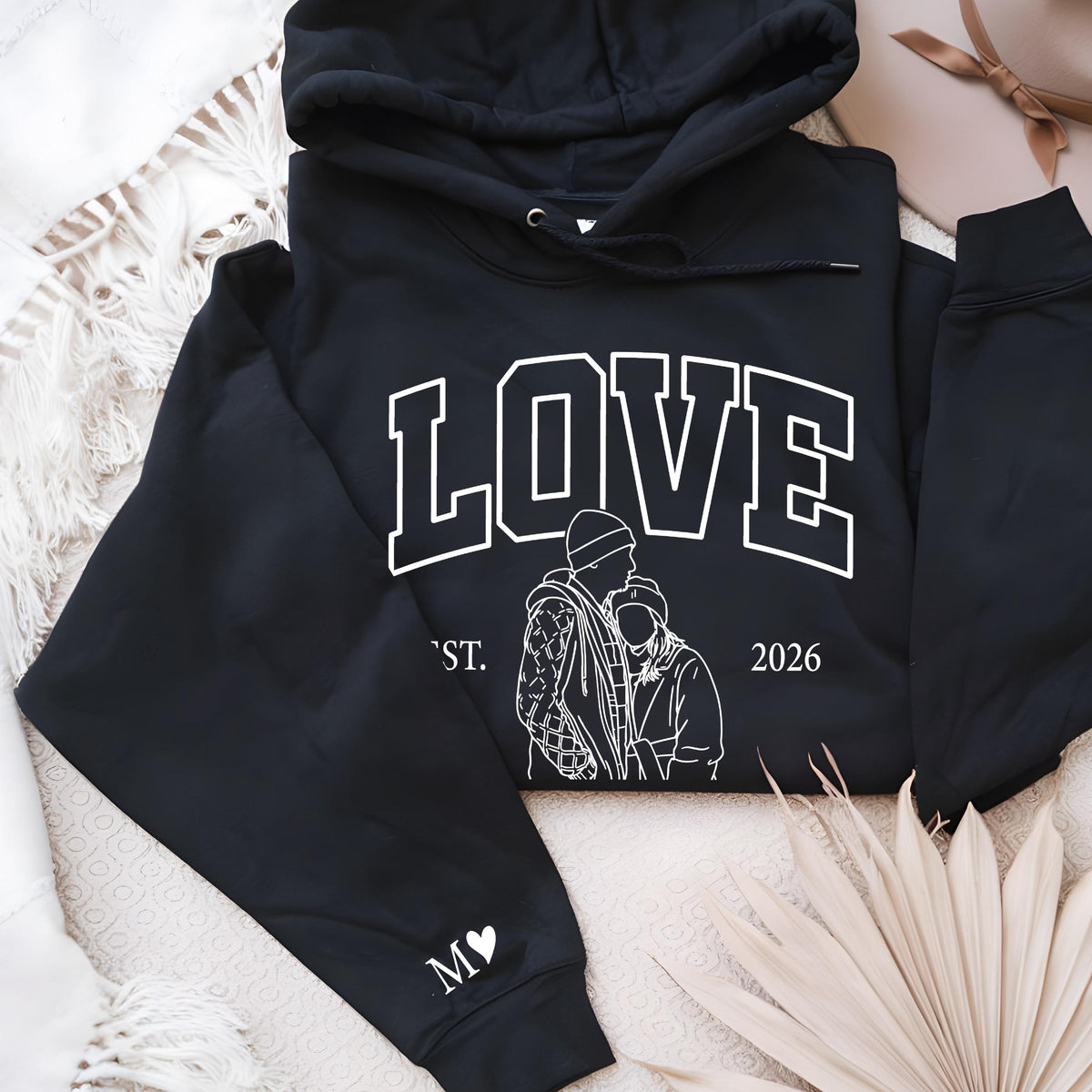 May include: Black hoodie with the word "LOVE" in white block letters. Below the word "LOVE" is a white line drawing of two people embracing. The year "2026" is printed on the right side. The left sleeve has a white heart and the letter "M".