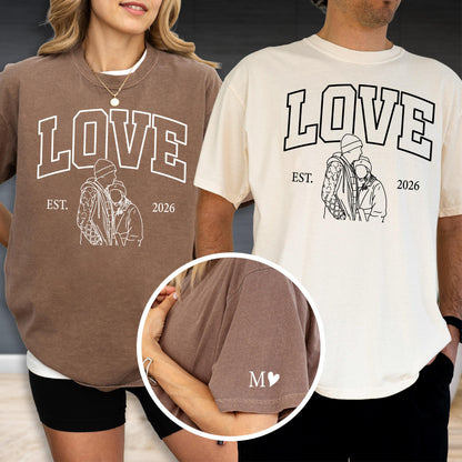 May include: Two t-shirts, one brown and one cream, each with the word "LOVE" in a bold, outlined font. Below the word is a line drawing of two people, with the text "EST. 2026". The brown shirt has an "M" with a heart on the sleeve.