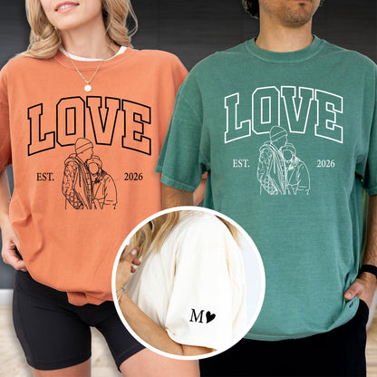 May include: Two matching t-shirts, one orange and one green, each with the word "LOVE" in bold letters. Below the word is a line drawing of two people embracing, with "EST. 2026" underneath. A third shirt is cream colored with the letter "M" and a heart.