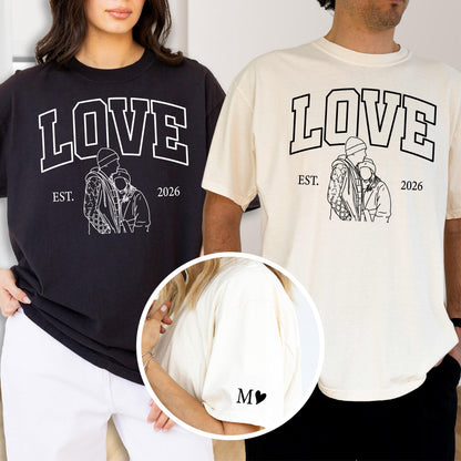 May include: Black and cream-colored t-shirts with the word "LOVE" in a bold, white, collegiate-style font. Below the word "LOVE" is a line drawing of two people embracing, with the text "EST. 2026". The left shirt is black, and the right shirt is cream.