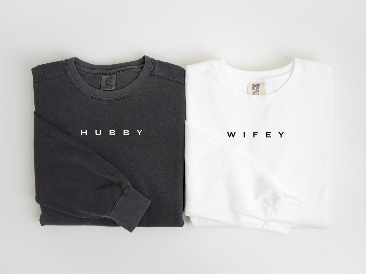 May include: Two long-sleeved sweatshirts, one charcoal gray and one white, are displayed side-by-side. The gray sweatshirt has the word "HUBBY" printed in white, while the white sweatshirt has "WIFEY" printed in black. Both are folded.