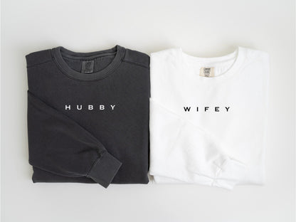 May include: Two long-sleeved sweatshirts, one charcoal gray and one white, are displayed side-by-side. The gray sweatshirt has the word "HUBBY" printed in white, while the white sweatshirt has "WIFEY" printed in black. Both are folded.