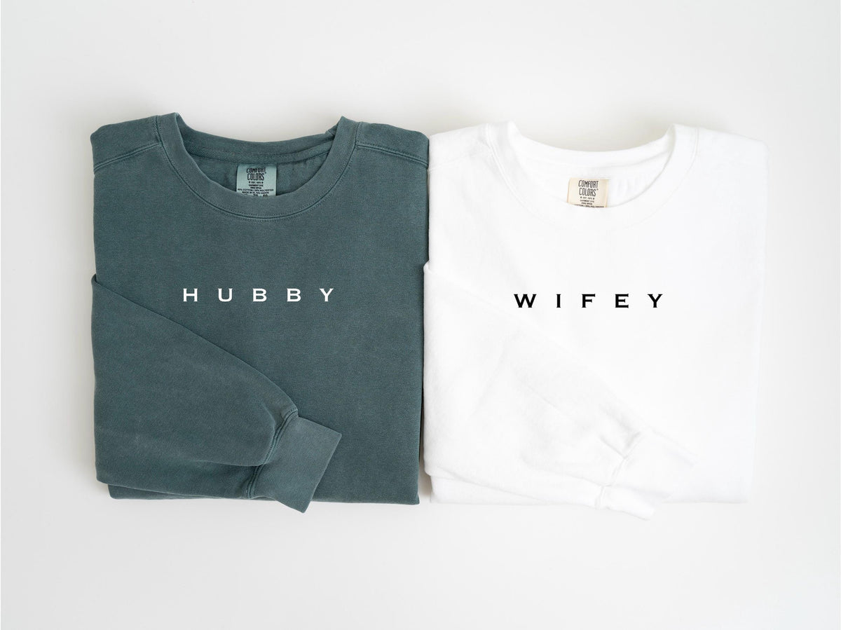 May include: Two long-sleeved shirts, one teal and one white, are displayed. The teal shirt has the word "HUBBY" printed on the front, while the white shirt has "WIFEY". Both shirts are folded neatly, showcasing a casual style.