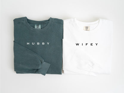 May include: Two long-sleeved shirts, one teal and one white, are displayed. The teal shirt has the word "HUBBY" printed on the front, while the white shirt has "WIFEY". Both shirts are folded neatly, showcasing a casual style.