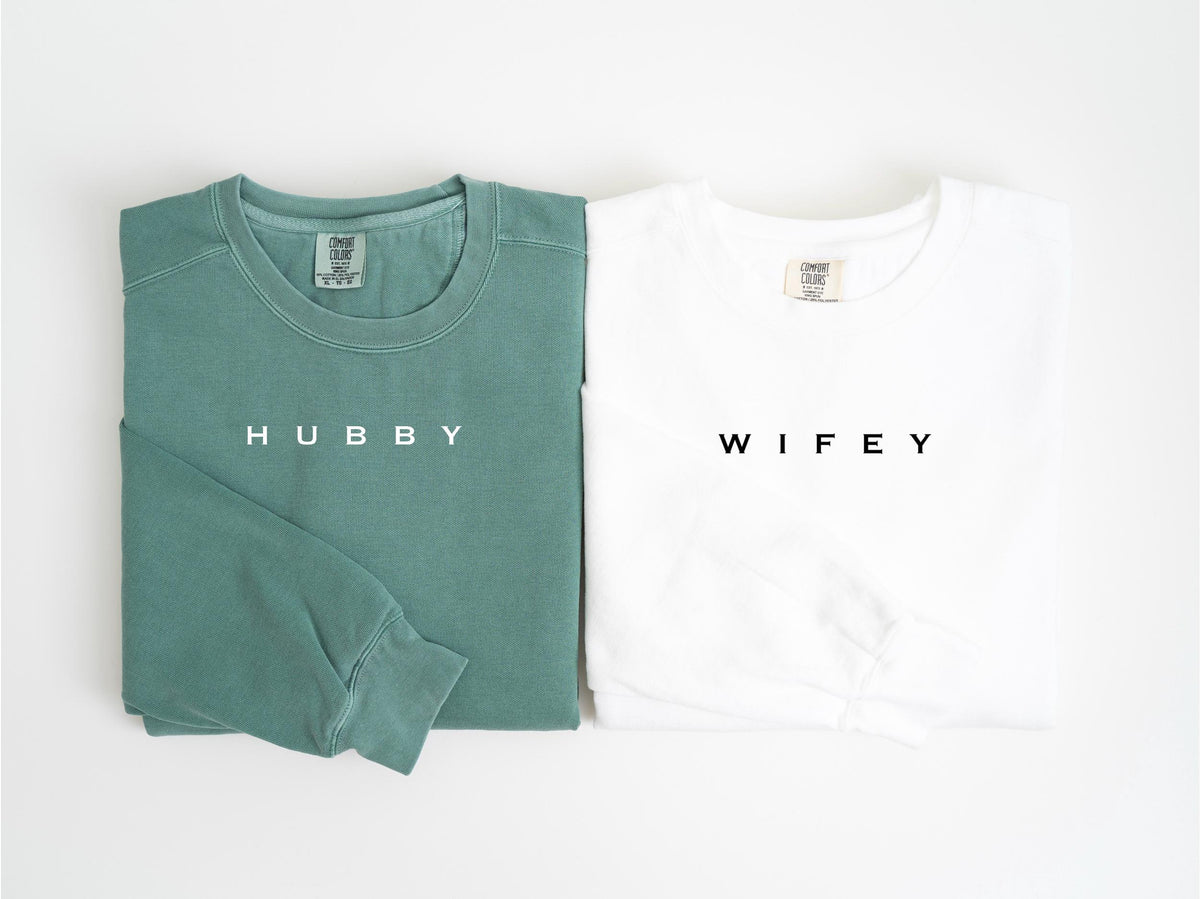 May include: Two long-sleeved sweatshirts, one teal and one white, are displayed side by side. The teal sweatshirt has the word "HUBBY" printed in white, while the white sweatshirt has "WIFEY" printed in black. Both have a crew neck.
