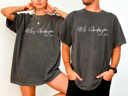 May include: Two matching charcoal gray t-shirts. One says "Mrs. Anderson EST. 2023" and the other says "Mr. Anderson EST. 2023" in white script. The shirts are worn by a couple, suggesting a wedding or anniversary theme.