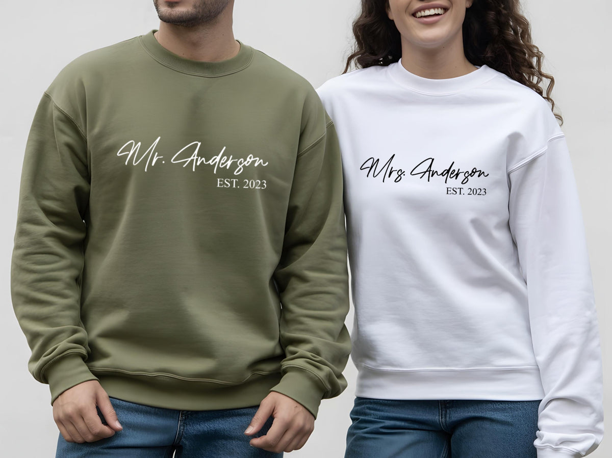 May include: Two matching sweatshirts, one olive green and one white. The olive green sweatshirt has "Mr. Anderson EST. 2023" in white script. The white sweatshirt has "Mrs. Anderson EST. 2023" in black script. Both are crew neck style.