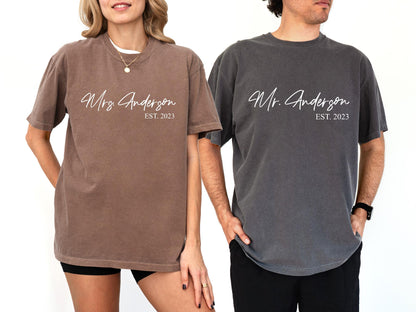 May include: Two matching t-shirts, one brown and one charcoal gray, each with the name "Anderson" in elegant script. The brown shirt reads "Mrs. Anderson EST. 2023", while the gray shirt says "Mr. Anderson EST. 2023".