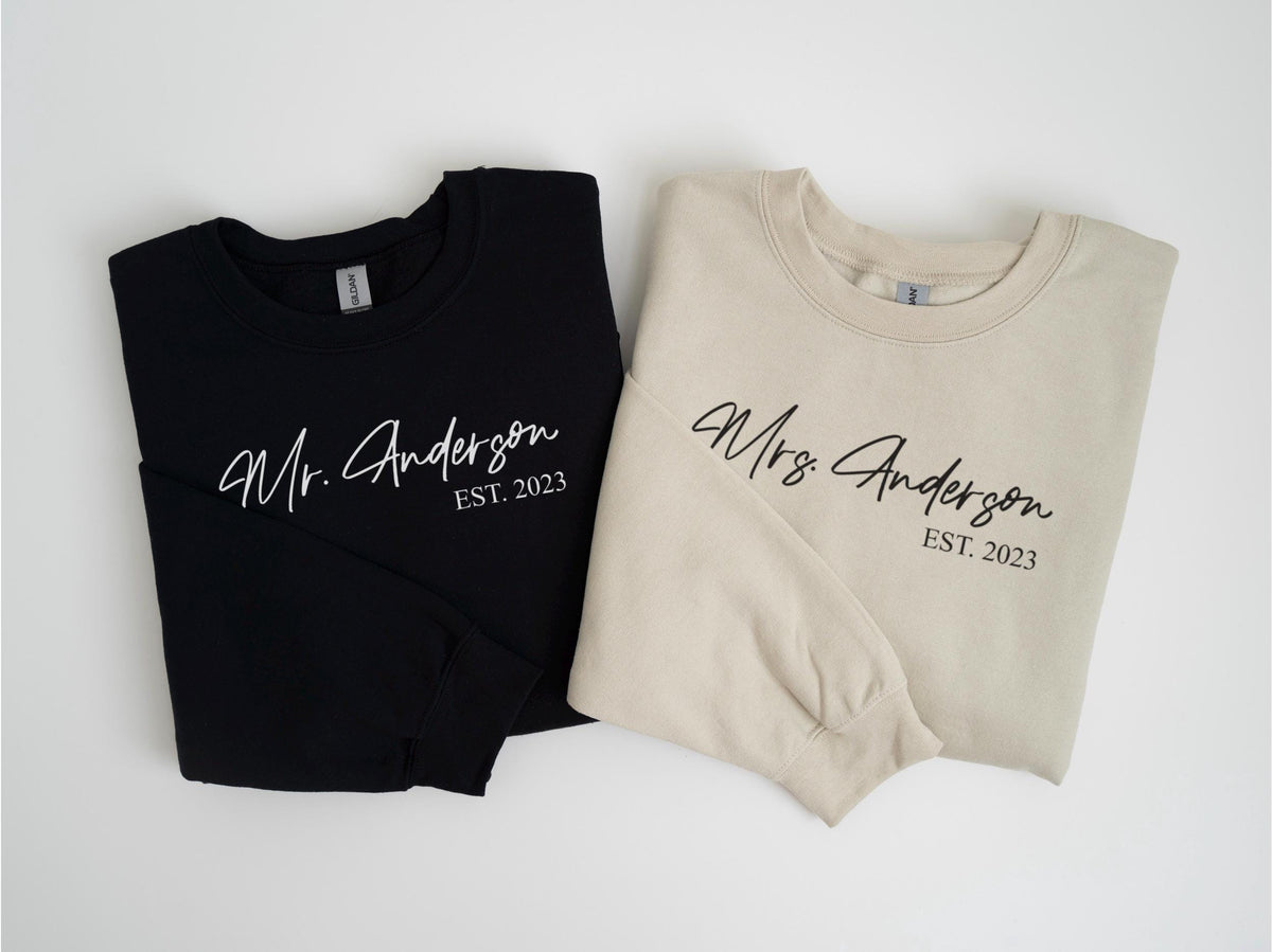 May include: Two crewneck sweatshirts, one black and one beige, with the names "Mr. Anderson" and "Mrs. Anderson" printed in a script font, along with "EST. 2023".